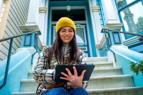 Young woman in a yellow beanie and plaid jacket sits on victorian steps in san francisco, smiling