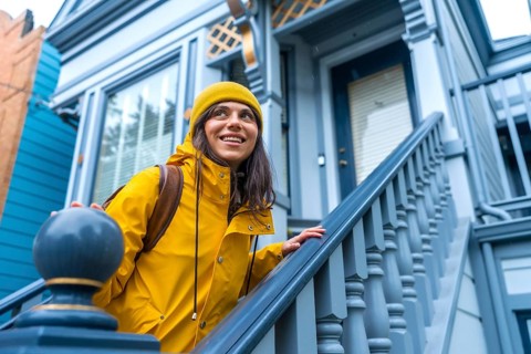 Woman wearing yellow raincoat and beanie smiling while ascending the exterior stairs of a classic
