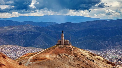 A hill with a statue above a city surrounded by mountains under a cloudy sky, The silver mines near