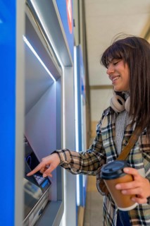 Smiling young woman engaging with an automated teller machine, holding a takeaway coffee cup,