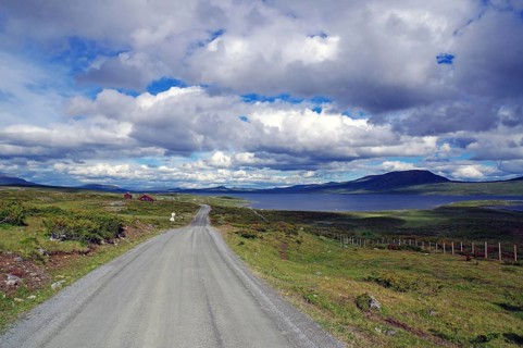 A country road leads through an open landscape with a view of a lake and distant mountains, Fjell,