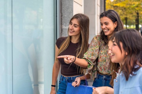 Three friends window shopping in the city, smiling and pointing at a store display, carrying bags