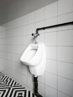 A urinal is mounted on a wall in a restroom. The tiles are white, and the floor has a pattern of