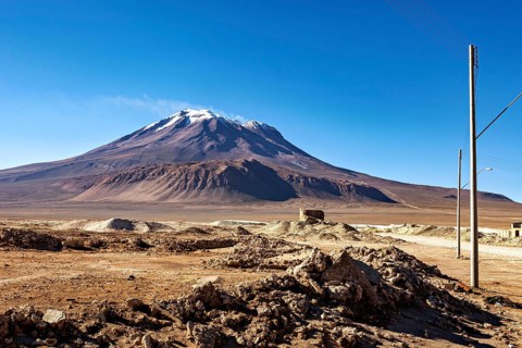 Large, snow-covered mountain above a barren, rocky landscape under clear skies, the town of Ollagüe
