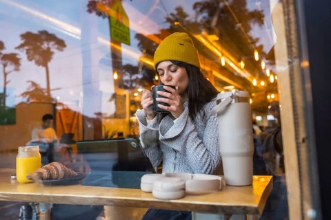 Young woman wearing a beanie enjoying a warm drink and breakfast at a wooden counter inside a
