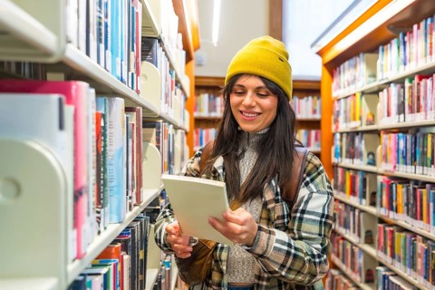 Female student wearing a casual outfit and a yellow beanie browsing books on shelves, smiling while