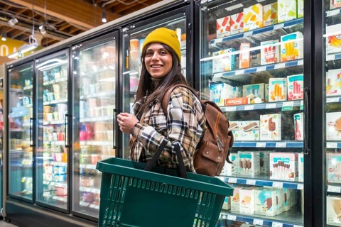 Young woman shopping for groceries in a supermarket aisle, smiling and holding a shopping basket