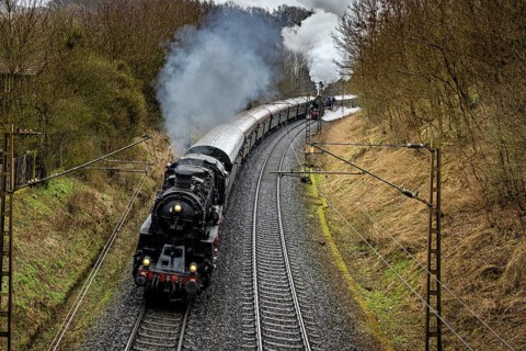 A steaming locomotive pulls a long train through a wooded bend, historic steam locomotive journey