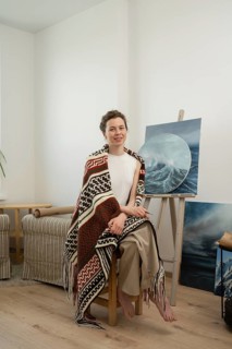 An artist sits barefoot in a cozy home studio, draped in a colorful patterned blanket. She is
