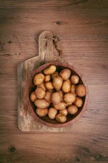 A collection of small, golden-brown potatoes fills a rustic wooden bowl placed on a textured wooden