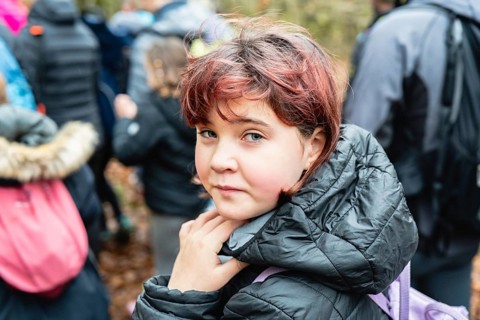 A young girl with a thoughtful expression, wearing a black jacket, stands among a group of people