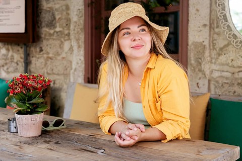 A young woman in a vibrant yellow shirt and sun hat sits at a rustic cafe table with a potted