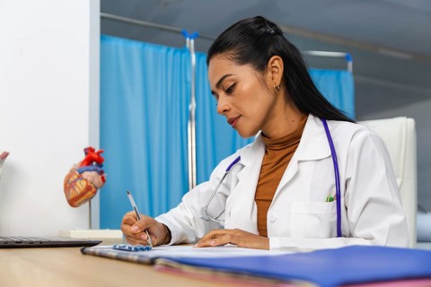 A dedicated doctor in a white coat writes notes at a desk in a medical office. A heart model is