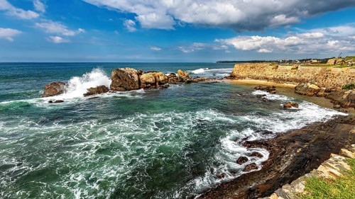Coastal landscape with waves crashing against rocks under a cloudy blue sky, The rocky coast in the
