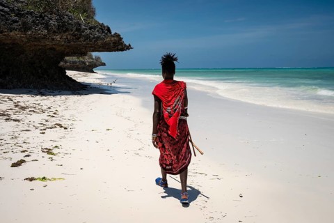 A Maasai man in traditional red attire walks away from the camera along a pristine white sand