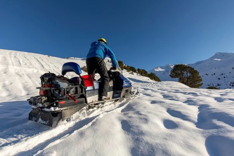 A snowmobiler in blue jacket navigates snowy terrain under a clear winter sky. The sun casts long