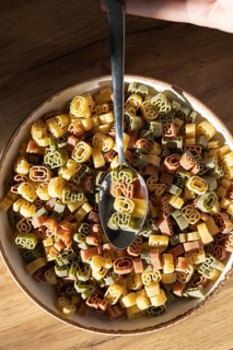 Top view of a bowl filled with multicolored, number shaped pasta being served with a spoon. The