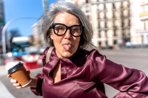 Playful woman expressing humor with her tongue out, holding a coffee cup and wearing eyeglasses on