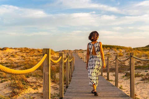 Woman walking on a wooden boardwalk, surrounded by golden sand dunes and dry grass, heading towards