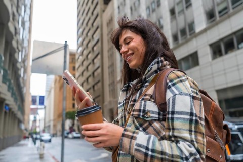 Young woman smiling while interacting with her smartphone and holding a coffee cup, walking