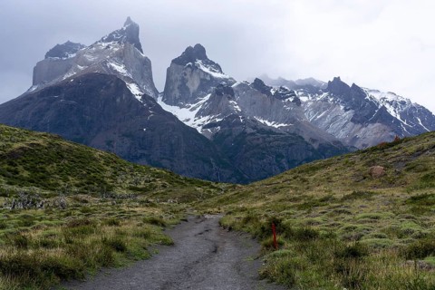 Cuernos del Paine, snow-capped granite peaks under cloudy sky, hiking trail with red marking leads