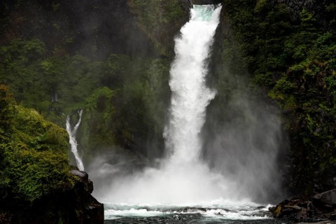 Waterfall falls over dark rock face in Gumpe, surrounded by thick rainforest with clouds of fog,
