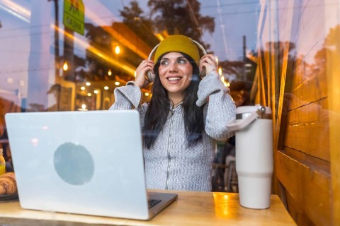 Young woman confidently adjusting headphones while sitting at a coffee shop window with a laptop