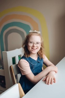 A joyful young girl with Down syndrome sits at a table in a colorful, playful room. She is smiling
