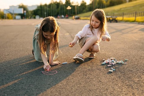 Two children enjoy drawing with colorful chalk on an asphalt surface during golden hour. The scene