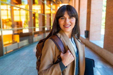 Female student standing outdoors on a modern university campus, carrying a backpack and a folder,