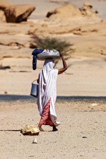 A woman carries loads on her head and a bucket in her hand in a remote desert landscape, woman