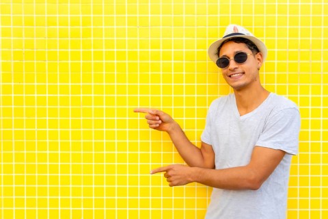 Stylish tourist pointing both index fingers at yellow tiled wall, wearing a hat and sunglasses
