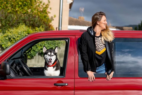 A smiling woman leans out of a red truck window while a friendly husky dog sits in the driver's