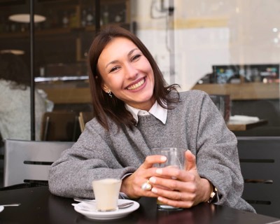 A woman in a gray sweater sits at an outdoor bar table, smiling warmly. She holds a glass, with a