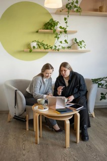 Two women engage in conversation in a cozy cafe surrounded by plants. They are working on a tablet,