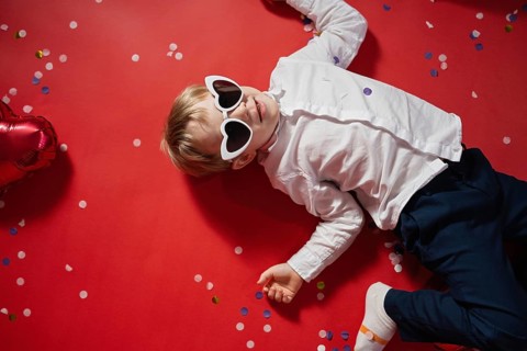 A young boy in heart-shaped sunglasses lies on a red background scattered with colorful confetti