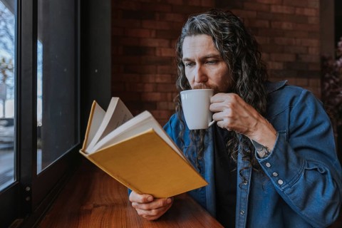 A man enjoys a tranquil moment in a cafe, sipping coffee while engrossed in a book by a window. The