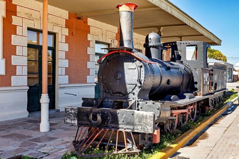 Old steam locomotive stands on tracks next to a historic railway station with brick architecture,