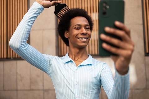 A non-binary individual with an afro hairstyle confidently takes a selfie outdoors. They are