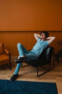 A young man with curly hair relaxes comfortably in a modern chair. His casual attire complements