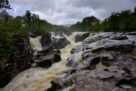 A river with several waterfalls flows through a rocky landscape surrounded by forest under cloudy