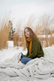 A woman sits on a blanket in a snowy park, surrounded by tall grasses. She wears a green sweater