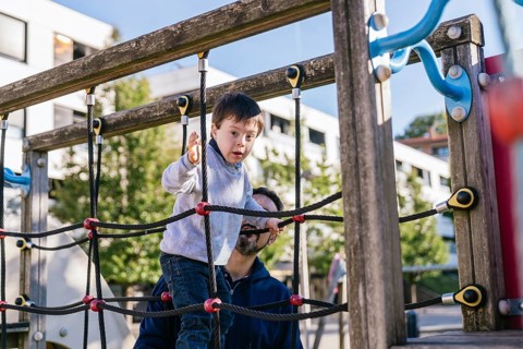 A child with Down syndrome explores a playground climber, supported by his father. The scene is