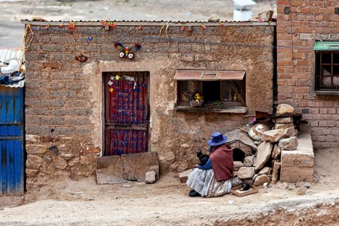 A woman in traditional dress sits on a floor in front of a brick wall with a decorated door, a