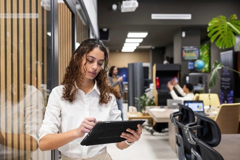 A professional woman in a white shirt uses a tablet in a stylish, modern office. Colleagues work