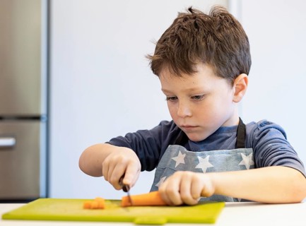 A young boy wearing a star patterned apron focuses intently on cutting a carrot with a small knife