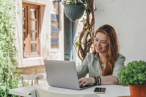 A woman working on a laptop at an outdoor table, surrounded by greenery. She is focused and