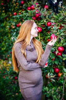 A beautiful pregnant woman with red hair stands in an apple orchard, surrounded by lush green