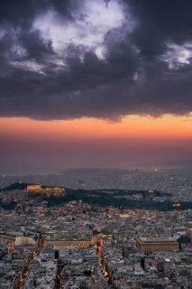 A dramatic sunset over Athens, Greece, with the Parthenon and Acropolis illuminated on the hilltop.