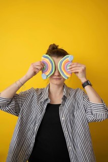 A person holds colorful clay rainbow shapes over their eyes, standing before a bright yellow